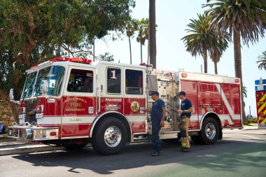 İtfaiyeciler, palmiye ağaçlarıyla dolu bir caddeye park edilmiş kırmızı bir itfaiye arabasının yanında duruyorlar. Şehir ortamında yanıp sönen ışıkları olan acil müdahale aracı. Kamu güvenliği, acil durum hizmetleri, itfaiyeciler ve şehir hayatı kavramı.