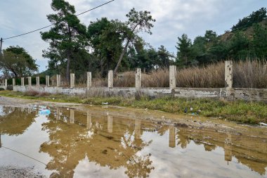 Terk edilmiş beton çitler, çamurlu kırsal bir yol boyunca, ağaçları yansıtan su birikintileriyle ve bulutlu gökyüzüyle vahşi otlar ve çam ormanlarıyla çevrili..