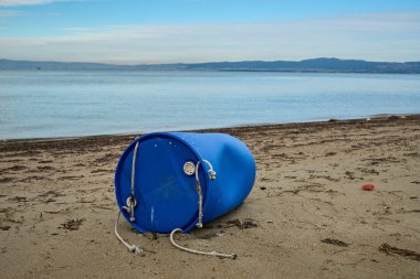 Mavi plastik varil kumlu bir plajda kıyıya vurmuş, deniz kirliliğini ve çevresel etkiyi simgeliyor, sakin deniz ve açık gökyüzü altındaki uzak tepeler.