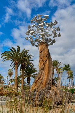 Mavi gökyüzünün altında palmiye ağaçlarıyla çevrili ahşap bir gövdeye monte edilmiş modern soyut metal heykel. Çağdaş kamu sanatı, açık hava kurulumu, seyahat ve kentsel konsept.