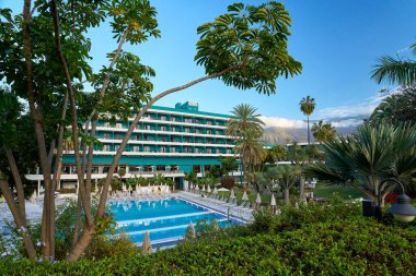 Etrafı tropik palmiye ağaçları, egzotik bitkiler ve yeşil bahçelerle çevrili büyük bir açık yüzme havuzu olan lüks bir otel. Açık mavi gökyüzü altındaki modern otel binası rahatlatıcı bir tatil atmosferi yaratıyor, seyahat için ideal.