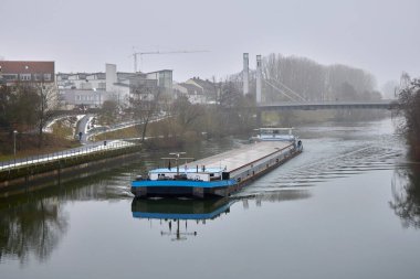 Şehir manzarasında sakin bir nehirde uzun bir yük mavnası geziniyor. Köprü, nehir kenarındaki binalar ve kış atmosferiyle iç su yolu taşımacılığı.