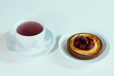 Bir fincan çay ve beyaz tabakta vişneli peynirli tart. Minimum temiz arkaplan, hafif ışık, basit tatlı ve içecek kompozisyonu.