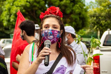 Bolsonaro 'nun itham edilmesini talep eden bir gösteri. Saçında çiçekler olan, maskeli bir kadın, protesto etmek için mikrofona konuşuyor..