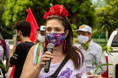 Bolsonaro 'nun itham edilmesini talep eden bir gösteri. Saçında çiçekler olan, maskeli bir kadın, protesto etmek için mikrofona konuşuyor..