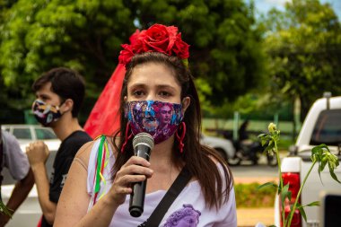 Bolsonaro 'nun itham edilmesini talep eden bir gösteri. Saçında çiçekler olan, maskeli bir kadın, protesto etmek için mikrofona konuşuyor..