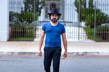  Şapkalı, mavi bluzlu ve siyah pantolonlu bir adam karşıdan karşıya geçiyor..