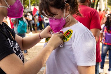 Diğer kızın bluzuna yapıştırıcı yapıştıran bir kız. Fotoğraf, Brezilya Başkanı Bolsonaro 'ya karşı yapılan bir protesto sırasında çekildi ve nüfusu aşılamak için aşı satın almada çeviklik talep ediliyor..