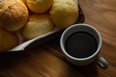 Bir fincan kahve ve peynirli çörek. Peynir ekmeği dolu küçük bir hasır sepet ve tahtadan bir yüzey üzerinde kahve olan beyaz bir fincan. Kahvaltı..