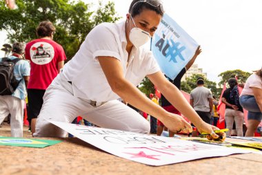 Kadın, koruyucu maske takıyor, gösteri için posterler yapıyor. Protesto; Brezilya Cumhurbaşkanı Bolsonaro 'ya karşı düzenlenen bir protesto sırasında çekilen fotoğraf ve nüfusu bağışık hale getirmek için aşı satın almada çeviklik istenmesi.