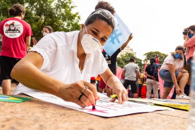 Kadın, koruyucu maske takıyor, gösteri için posterler yapıyor. Protesto; Brezilya Cumhurbaşkanı Bolsonaro 'ya karşı düzenlenen bir protesto sırasında çekilen fotoğraf ve nüfusu bağışık hale getirmek için aşı satın almada çeviklik istenmesi.