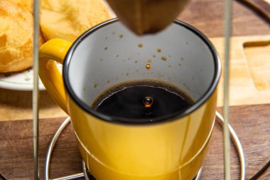 Altında sarı bir kupayla kumaş süzgecine kahve sıkıyor. Arka planda peynirli çörek tabağı. Kahvaltı hazırlıyorum. Brezilya 'dan tipik yemek..