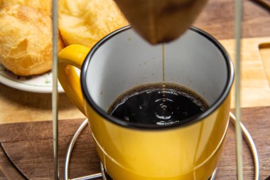 Altında sarı bir kupayla kumaş süzgecine kahve sıkıyor. Arka planda peynirli çörek tabağı. Kahvaltı hazırlıyorum. Brezilya 'dan tipik yemek..