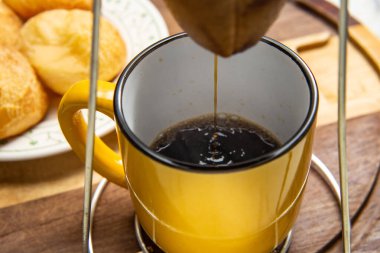 Altında sarı bir kupayla kumaş süzgecine kahve sıkıyor. Arka planda peynirli çörek tabağı. Kahvaltı hazırlıyorum. Brezilya 'dan tipik yemek..