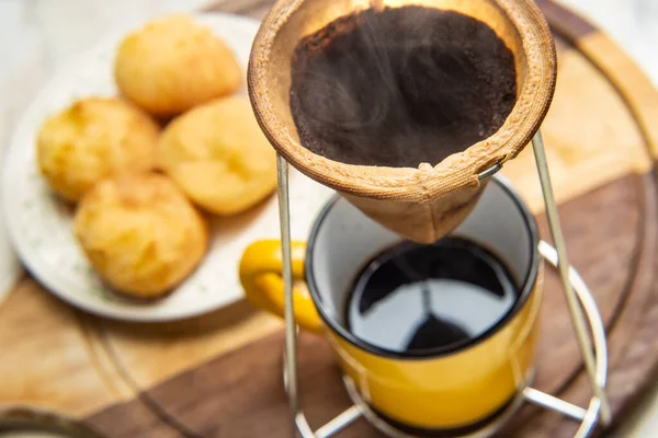 Altında sarı bir kupayla kumaş süzgecine kahve sıkıyor. Arka planda peynirli çörek tabağı. Kahvaltı hazırlıyorum. Brezilya 'dan tipik yemek..