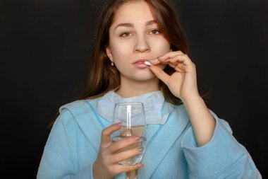 Siyah arka planda elinde bir hap ve bir bardak su tutan genç bir kadın.