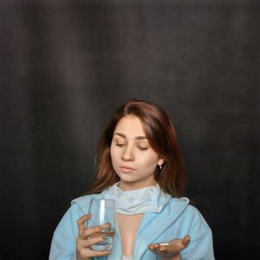 Siyah arka planda elinde bir hap ve bir bardak su tutan genç bir kadın.