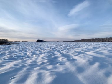 Mavi gökyüzünün altında, tüylü bulutlarla kaplı bir tarla. Kış manzarası mavi tonlarda ve arka planda ağaçlar olan kar yağışı yüzeyi.