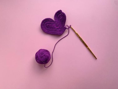 Sevgililer Günü temalı tığ işi. Fotoğrafta, bir kanca, bir leylak kalp, bir ip yumağı. Yukarıdan bir fotoğraf. Ürün yaratma kavramı. Pembe arka planda örülmüş bir kalp