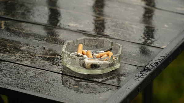 Wet ashtray with extinguished cigarette on the table after the rain. Bad habits concept. Ashtray for cigarettes, side view, copy space. n