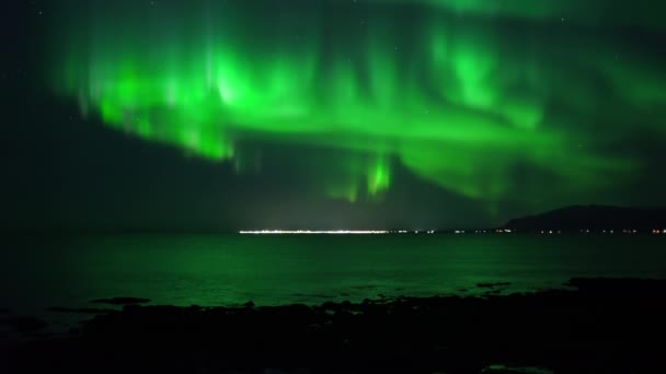 Real-time active aurora borealis reflecting in ocean over far town Iceland — Stock video