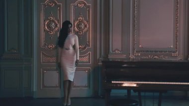 Uzun beyaz bir elbise mükemmel rakam uzun siyah saçlı seksi güzel kadın piyano de geçer klasik tarzı altın sıva oyun, zarif podyum başlar dekore edilmiş kapıdan uzak hareket ediyor
