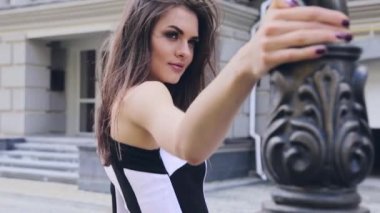 Genç güzel seksi kız gün flört saklandığı bir sütun arkasında sırasında sokakta yürüyen görünüyor ve rüzgarla geliştirme gülümsüyor uzun siyah saçlı siyah ve beyaz bir elbiseyle tek eliyle tutun