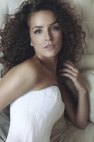 Portrait of curly dark-haired young beautiful girl in a white corset sitting on the couch in semi with upholstered headboard — Stock Photo, Image