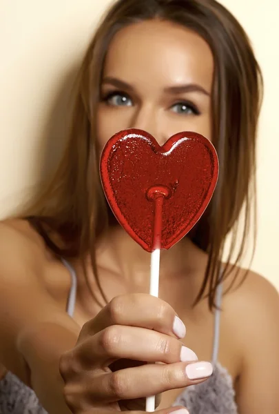 Hermosa joven con una bata de seda y encaje con correas delgadas con un corte y arco delante de las manos sosteniendo una piruleta en la forma de un día de San Valentín corazón rojo — Foto de Stock