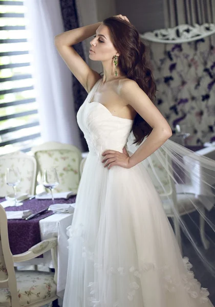 Beautiful young bride with long brown wavy hair in a lush white wedding dress of tulle, embroidered with beads corset, bow tied at the waist photographed in the interior of the restaurant — Stock Photo, Image