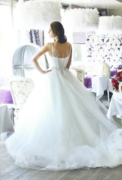 Bela noiva jovem com cabelo ondulado marrom longo em um vestido de noiva branco exuberante de tule, bordado com espartilho de contas, arco amarrado na cintura fotografada no interior do restaurante — Fotografia de Stock