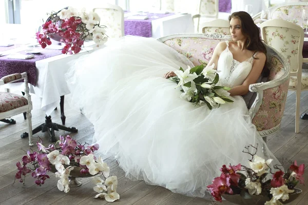 Hermosa joven novia con el pelo largo y ondulado marrón en un exuberante vestido de novia blanco de tul, bordado con corsé de cuentas, lazo atado a la cintura fotografiado en el interior del restaurante — Foto de Stock