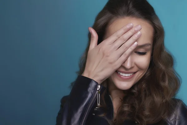 Portret van de krullend mooi verlegen meisje met glanzende witte tanden en donkere haren verbergen een oog en lacht op een blauwe achtergrond in lederen jas — Stockfoto