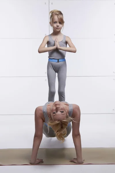 Mother and daughter doing yoga exercise, fitness, gym wearing the same comfortable tracksuits family sports, sports paired woman put her hands on the floor child standing on her abdomen girl balancing — Stock Photo, Image