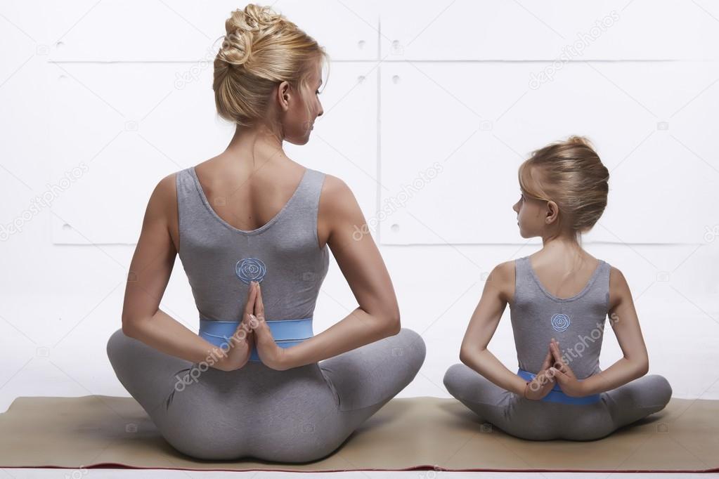 Mother and daughter doing yoga exercise, fitness, gym wearing the same comfortable tracksuits, family sports, sports paired siting back on relax pose and holding hand back they are on skinny suit