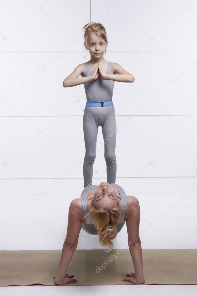 Mother and daughter doing yoga exercise, fitness, gym wearing the same comfortable tracksuits family sports, sports paired woman put her hands on the floor child standing on her abdomen girl balancing