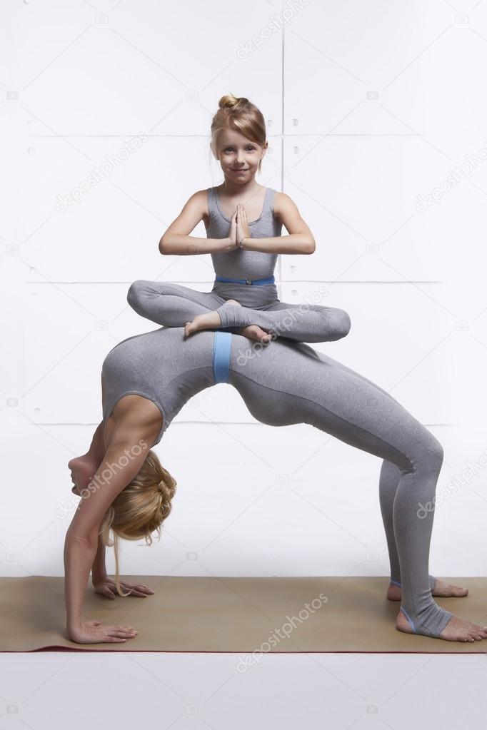 Mother and daughter doing yoga exercise, fitness gym wearing same woman standing in posture of bridge hands and feet resting on the floor arching his back girl sitting on her abdomen in lotus position