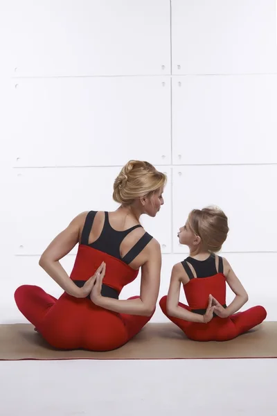 Mother and daughter doing yoga exercise, fitness, gym wearing the same comfortable tracksuits, family sports, sports paired siting back on relax pose and holding hand back they are on red skinny suit — Stock Photo, Image