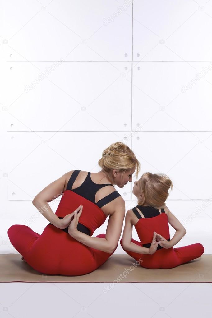 Mother and daughter doing yoga exercise, fitness, gym wearing the same comfortable tracksuits, family sports, sports paired siting back on relax pose and holding hand back they are on red skinny suit