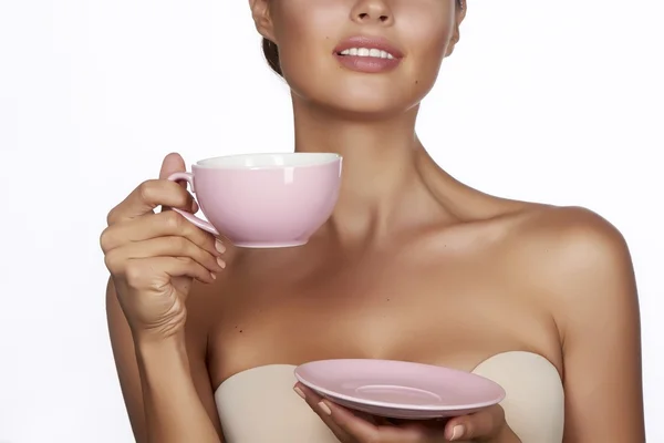 Young sexy beautiful woman with dark hair picked up holding a ceramic cup and saucer pale pink drink tea or coffee on a white background — Stock Photo, Image