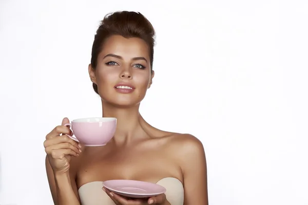 Young sexy beautiful woman with dark hair picked up holding a ceramic cup and saucer pale pink drink tea or coffee on a white background — Stock Photo, Image