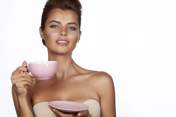 Young sexy beautiful woman with dark hair picked up holding a ceramic cup and saucer pale pink drink tea or coffee on a white background — Stock Photo, Image
