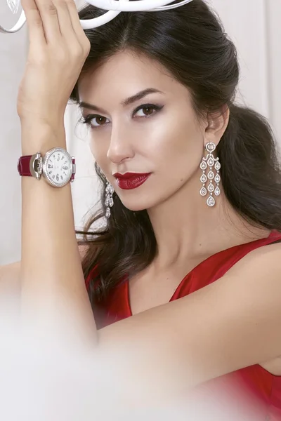 Hermosa mujer sexy morena Ojos marrones en un vestido rojo en lujosos pendientes con diamantes y relojes en la correa de cuero de Borgoña maquillaje de noche, lápiz labial rojo, levantó una mano —  Fotos de Stock