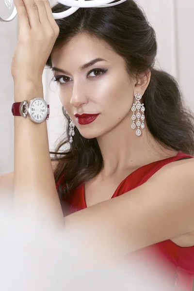 Hermosa mujer sexy morena Ojos marrones en un vestido rojo en lujosos pendientes con diamantes y relojes en la correa de cuero de Borgoña maquillaje de noche, lápiz labial rojo, levantó una mano —  Fotos de Stock