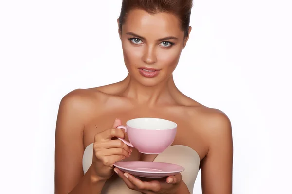 Young sexy beautiful woman with dark hair picked up holding a ceramic cup and saucer pale pink drink tea or coffee on a white background — Stock Photo, Image