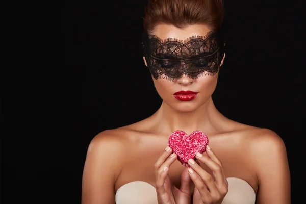 Young beautiful sexy woman with dark lace on eyes bare shoulders and neck, holding cake shape of heart to enjoy the taste and are dieting, feeling temptation, teeth passion sex red lips — Stock Photo, Image