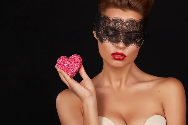 Young beautiful sexy woman with dark lace on eyes bare shoulders and neck, holding cake shape of heart to enjoy the taste and are dieting, feeling temptation, teeth passion sex red lips — Stock Photo, Image