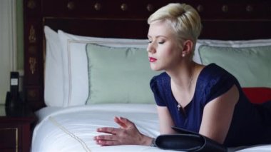 Lüks otel yatak odasında sıska kısa elbise takı güneş gözlüğü giyen güzel seksi sarışın iş kadını içki şarap toplantı dan önce dinlenmek moda trendi kırmızı ruj yastık çarşafları makyaj yapmak