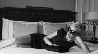 Lüks otel yatak odasında sıska kısa elbise takı güneş gözlüğü giyen güzel seksi sarışın iş kadını içki şarap toplantı dan önce dinlenmek moda trendi kırmızı ruj yastık çarşafları makyaj yapmak