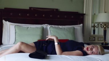 Lüks otel yatak odasında sıska kısa elbise takı güneş gözlüğü giyen güzel seksi sarışın iş kadını içki şarap toplantı dan önce dinlenmek moda trendi kırmızı ruj yastık çarşafları makyaj yapmak